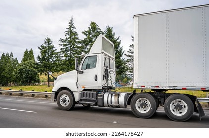 Big rig white industrial diesel day cab semi truck with roof spoiler and dry van semi trailer transporting commercial cargo load driving on the wide multiline highway road with trees on the side