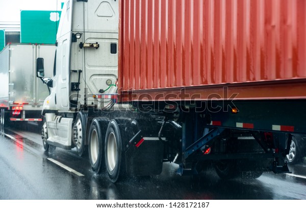 Big rig white long haul semi truck transporting\
commercial cargo in red container running with turned on headlights\
on rain dusted wet raining highway driving with another semi trucks\
heavy traffic 