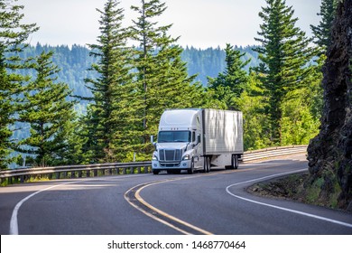 Big rig white powerful American bonnet long haul semi truck transporting commercial cargo in refrigerated semi trailer moving uphill on winding road with a green trees and safety fence on the side