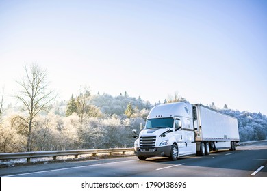 Big rig white technological improved long haul semi truck with refrigerated semi trailer for transportation of perishable and frozen food going on the winter road with snowy frost trees on the hill