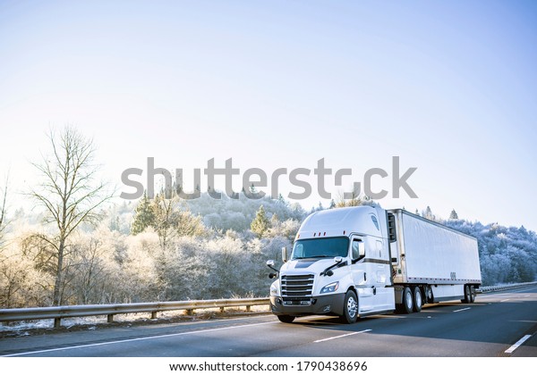 Big rig white technological improved long haul semi\
truck with refrigerated semi trailer for transportation of\
perishable and frozen food going on the winter road with snowy\
frost trees on the hill