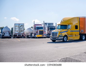 Big Rig Yellow Long Haul Semi Truck With Semi Trailer Running On The Truck Stop Parking Lot Past The Rest Of The Semi Trucks Standing In Row And Waiting For The Continuation Of The Delivery Route