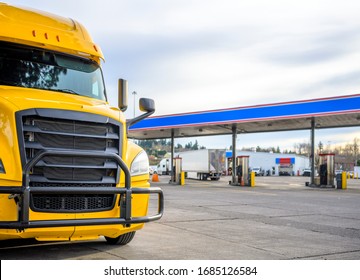 Big Rig Yellow Long Haul Industrial Diesel Semi Truck With Grille Guard Standing In Row With Another Semi Trucks And Semi Trailers On Truck Stop Parking Lot With Fuel Station For Refueling