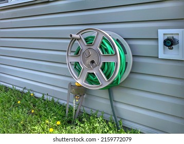 A Big Rolled Green Water Hose Attach To Metal Siding Wall With The Automatic Lawn Sprayer At The Edge, Next To The Outdoor Water Tap In The Backyard Of The House