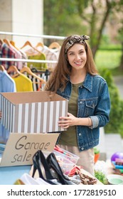 Big Sale. Pretty Happy Woman Standing With A Box While Organizing A Garage Sale