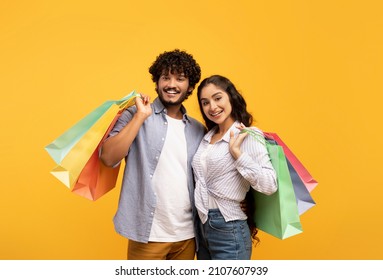 Big sale and shopping with pleasure. Happy young indian couple holding many colored packages in hands, posing on yellow background, studio shot, free space