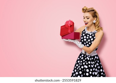 Big sales discounts, rebates offers ad concept - portrait image of happy smiling beautiful woman in pin up black dress with polka dot, white gloves, hold gift box, isolated rose pink background - Powered by Shutterstock