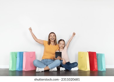 Big Sales. Happy Mother And Little Daughter With Digital Tablet And Lots Shopping Bags Sitting On Floor Near White Wall And Shaking Fists, Cheerful Mom And Child Enjoying Internet Purchases - Shutterstock ID 2311020681