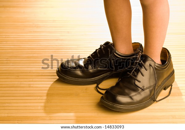 Big shoes to fill,\
child\'s feet in large grown-up black shoes, on backlit wood floor,\
playing dress-up