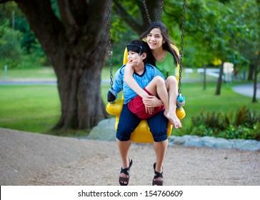 Big Sister Holding Disabled Brother On Special Needs Swing At Playground In Park. Child Has Cerebral Palsy.