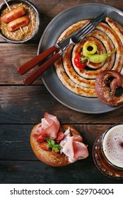 Big Spiral Fried Sausage, Meat Snacks Wienerwurst, Ham, Marinated Chili Peppers Served In Salted Pretzels And Plate With Glass Of Lager Beer And Mustard Over Dark Wooden Background. Top View