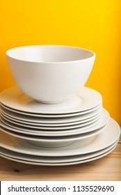 Big Stack Of Different White Plates, Bowls, Isoalted Yellow Background, One Above The Another. Wooden Table, Vertical Image Copy Space.