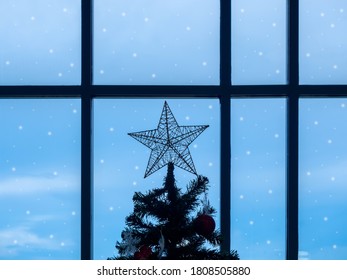 Big Star On Top Of Dark Christmas Tree Near Window Glass With Snow On Blue Sky Background In Winter Season, Lonely Feeling.