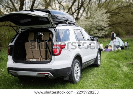 Big SUV car with open trunk and roof rack box against happy young family having fun and enjoying outdoor on picnic blanket at garden spring park.