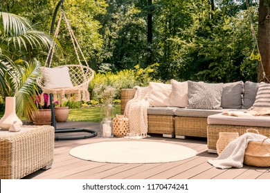 A big terrace with a comfortable leisure sofa with cushions, a table and a string swing in a green garden during sunny vacation.