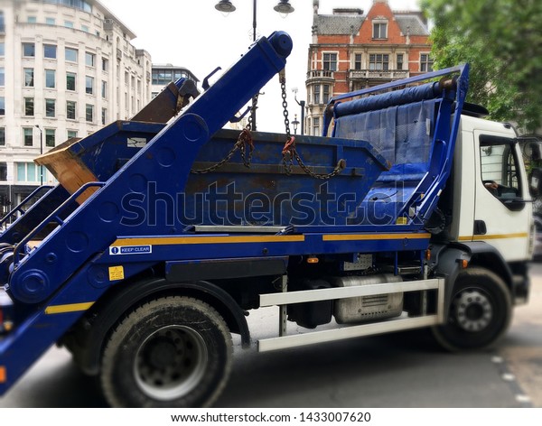 Big truck loaded with blue industrial skip.
Selective focus on the metal bin on back of the vehicle with blurry
surrounding area of truck, wheels, buildings as space to add text
for background use.