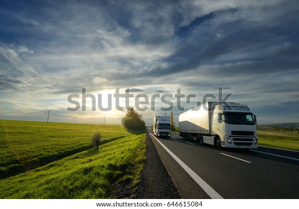 Big truck overtaking a small\
truck on a road in a rural landscape at sunset with dramatic\
clouds