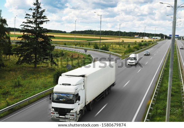 A big truck and white trailer with space for text and\
other cars driving fast on the countryside road against a cloudy\
sky