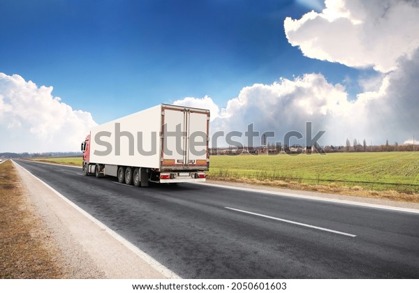 Big truck with a white trailer with a space for\
text on a countryside road with a green field against a blue sky\
with clouds