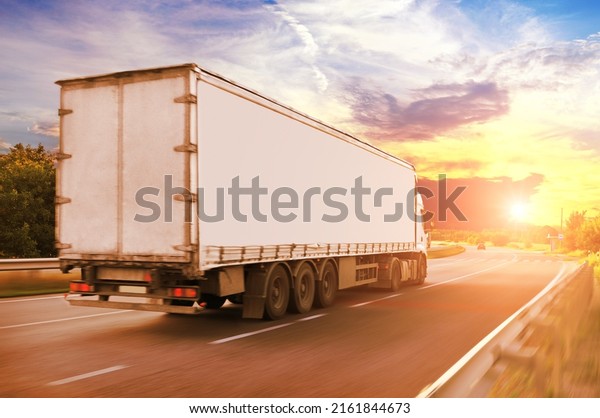 Big\
truck with a white trailer with space for text driving fast on a\
countryside road against an evening sky with a\
sunset