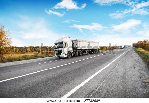 Big trucks and white trailers and
cars on the countryside road against blue sky with
clouds