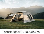 A big white camping tent with camping gears in a green grass with mountains and blue sky with white clouds in the background. Taken during golden hour