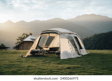 A big white camping tent with camping gears in a green grass with mountains and blue sky with white clouds in the background. Taken during golden hour