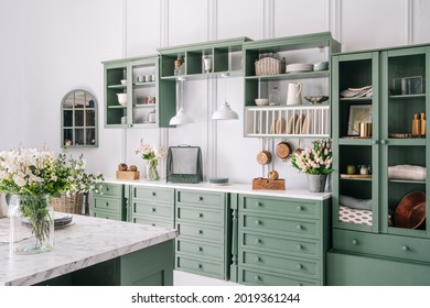 Big white kitchen with green furniture, convinient and well decorated house enviroment, cupboard with glass doors, marble countertop and jars filled with flowers, lots of drawers