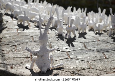 Big white rabbit statue made of plaster back view, outdoor art exhibition, artificial strange hare on city street. Handmade decorative bunny in front of flock of small rabbits, Easter urban decor