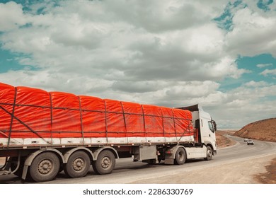 A big white trailer carries a heavy red load on a cloudy day on a mountain road. A truck with a cloudy sky background. A truck carries cargo. - Shutterstock ID 2286330769