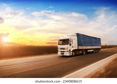 Big white truck with a blue trailer with blank space for text on a countryside road in motion against an evening  sky with a sunset