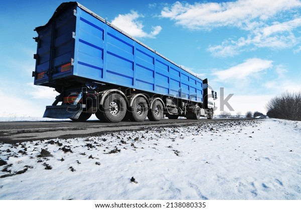 Big white truck and a blue trailer with space for\
text on a winter countryside road with snow against a blue sky with\
clouds