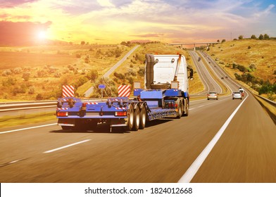 A Big White Truck With A Heavy Equipment Flatbed Trailer With Other Cars On A Countryside Road In Motion Against A Night Sky With A Sunet