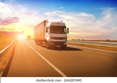 A big white truck with a red trailer and other cars on the countryside road in motion against a night sky with a sunset