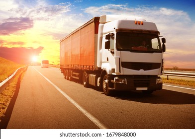 A Big White Truck And A Red Trailer With Other Vehicles On The Countryside Road Against A Night Sky With A Sunset