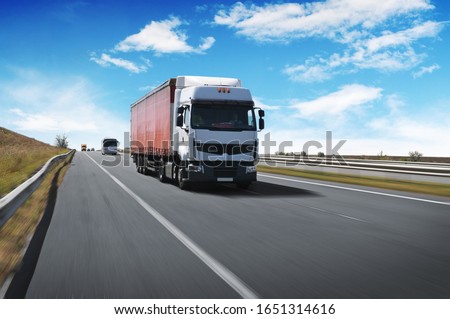 A big white truck with a red trailer and other cars on the countryside road in motion against a blue sky with clouds