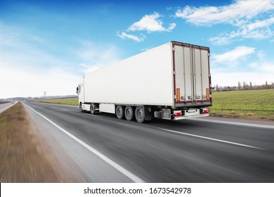 A Big White Truck And A Trailer With Space For Text On The Countryside Road In Motion Against A Blue Sky With Clouds