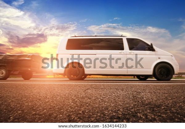 Big white van with a small\
trailer on a countryside road against a night sky with a\
sunset