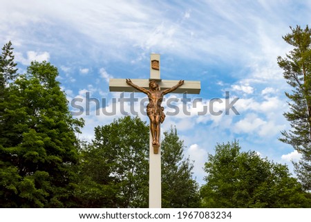 Big white wooden crucifix of Jesus Christ outdoors in a cemetery - bronze sculpture hit by sunlight against a blue sky with white clouds and green trees