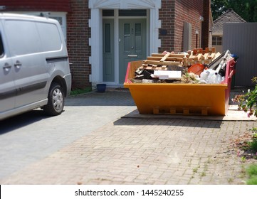 Big Yellow Industrial Skip In Front Of The House, One Side Of Frame. Focus On Full Metal Bin With Blurry Surrounding Area As Space To Add Text On Driveway Floor, White Van, Brick House In Background. 