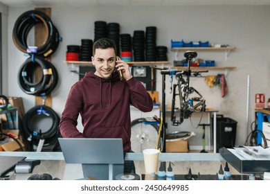 The bike mechanic is answering the phone with a smile, looking at the camera with a laptop in front - Powered by Shutterstock