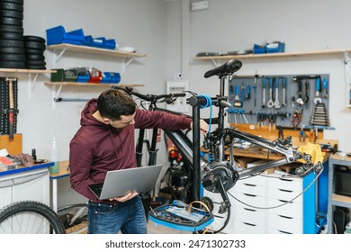 The bike mechanic is manipulating and repairing an electric mountain bike while holding a laptop - Powered by Shutterstock