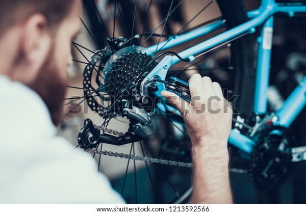 Bike Mechanic\
Repairs Bicycle in Workshop. Closeup Portrait of Young Blurred Man\
Examines and Fixes Modern Cycle Transmission System. Bike\
Maintenance and Sport Shop\
Concept