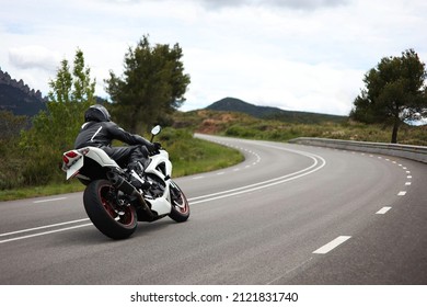 Biker in a black uniform driving a white sports motorcycle on a curvy road in the middle of a green meadow with trees and with mountains in the background on a cloudy day