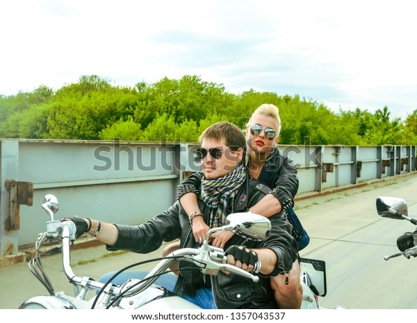 Biker Couple
with motorcycle Chopper style Man and woman ride with high
speed.girl wear leather jacket and sunglasses against park
background Gang of groups of armed people.
