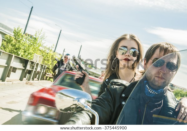 Biker Couple with motorcycle Chopper style Man and\
woman ride with high speed Cute girl wear black leather jacket and\
stylish sunglasses against urban background Gang of groups of armed\
people