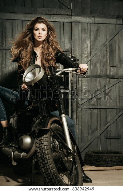 ビンテージバイクに乗った革のジャケットを着た 金髪のバイカー 女の子 美しい女性 バイク グレイの木の背景にガレージ バイク 趣味 ライフスタイル の写真素材 今すぐ編集