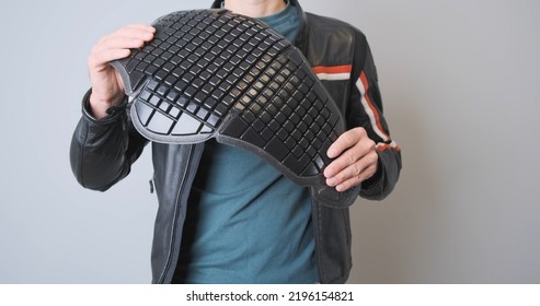 Biker In A Leather Black Sports Motorcycle Jacket, Holds A Plastic Back Protection In His Hands, Demonstrates It, Squeezes It. Front View, Close-up, Indoors.