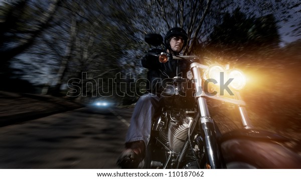 Biker
man riding his motorbike fast with motion blur and lens flare at
night while being chased by a car in the
distance.