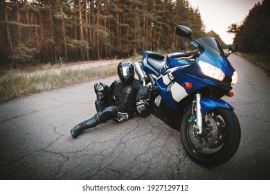 Biker in a protective suit and a black helmet sits next to his motorcycle on the road. Motorcyclist resting near a motorcycle